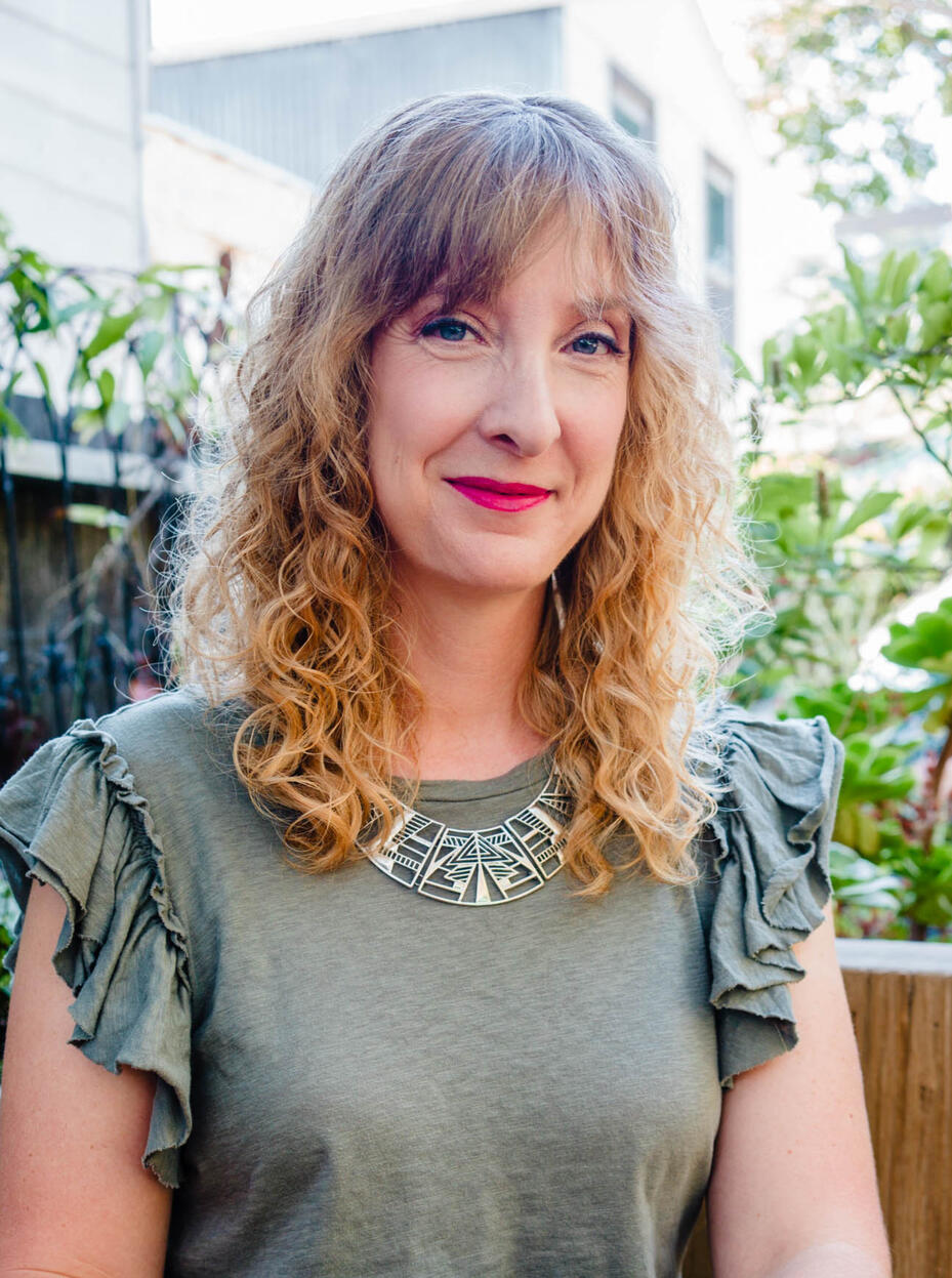 woman with dishwater blonde wavy hair smiles and looks directly at the camera at a table outside with trees in the background