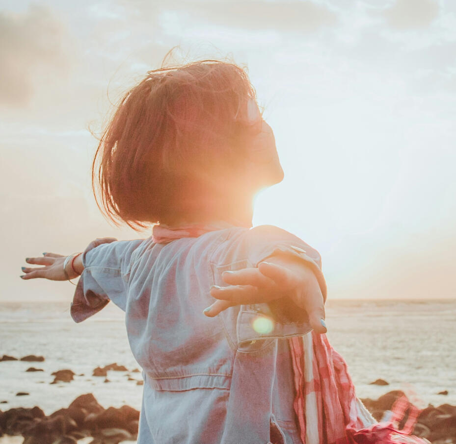 muted tones, woman faces sunset over the ocean on a rocky beach with hands outstretched. She's faced away from us.